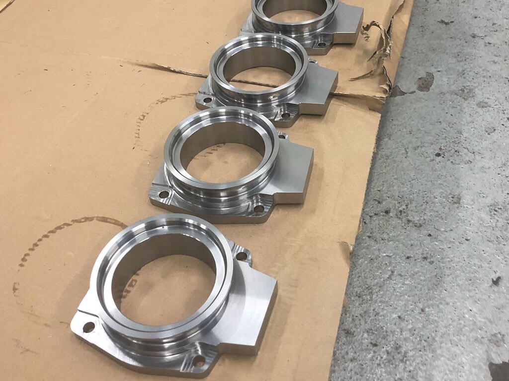 Precision-machined metal parts with a cylindrical shape and flanged base, arranged in a row on cardboard, showing polished surfaces.